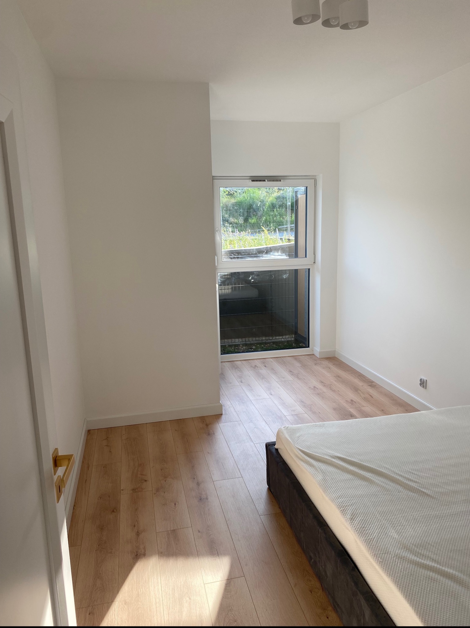 Jasna sypialnia po remoncie: parkiet, białe ściany i okno z widokiem na zieleń. Minimalistyczny wystrój, widoczny fragment łóżka i drzwi z złotą klamką.