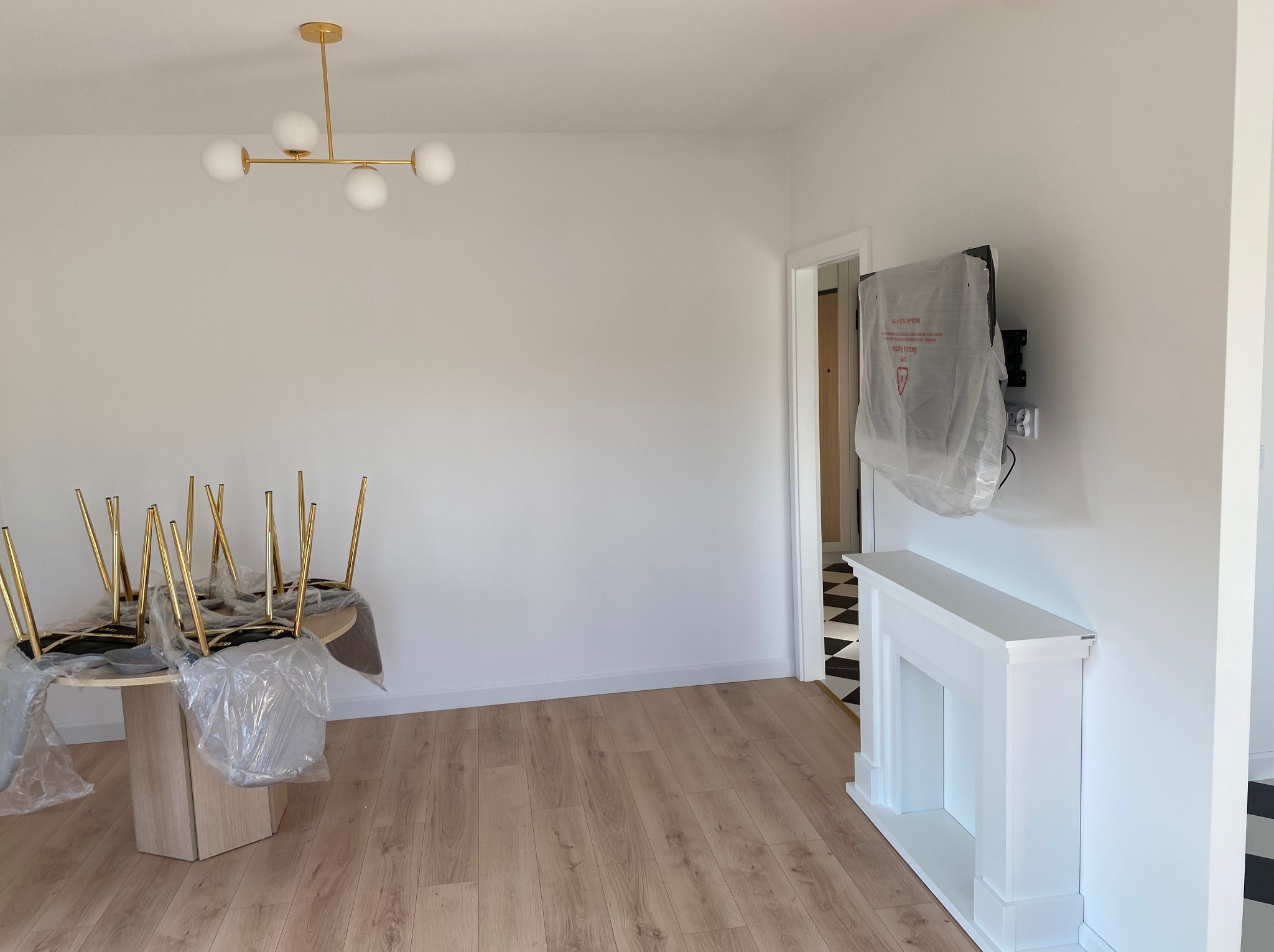 Jasne wnętrze po remoncie: panele podłogowe, białe ściany, ozdobny kominek, nowoczesna lampa. Krzesła i telewizor zabezpieczone folią.