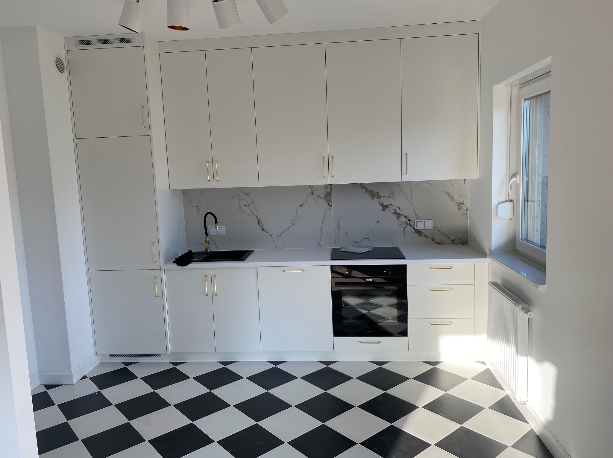 Biała kuchnia z czarnym blatem i złotymi uchwytami, wzorzysty panel ścienny imitujący marmur, czarno-biała podłoga w szachownicę, nowoczesny design.