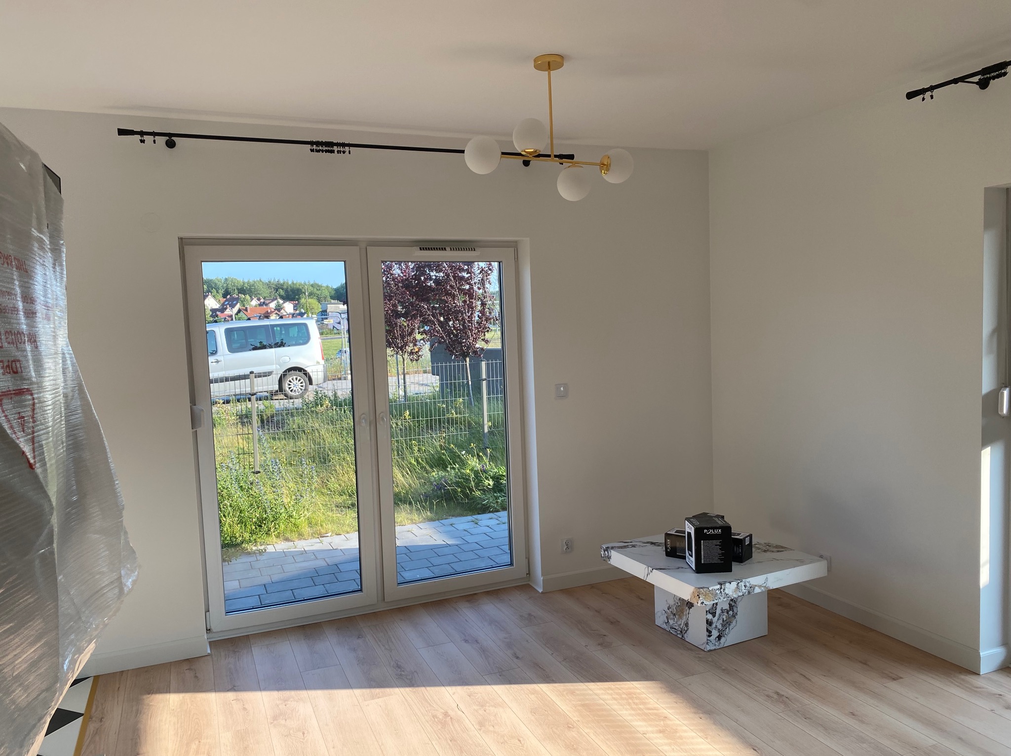 Jasny pokój po remoncie z panelami podłogowymi, marmurowym stolikiem i widokiem na zieleń przez duże okno balkonowe. Nowoczesna lampa sufitowa.