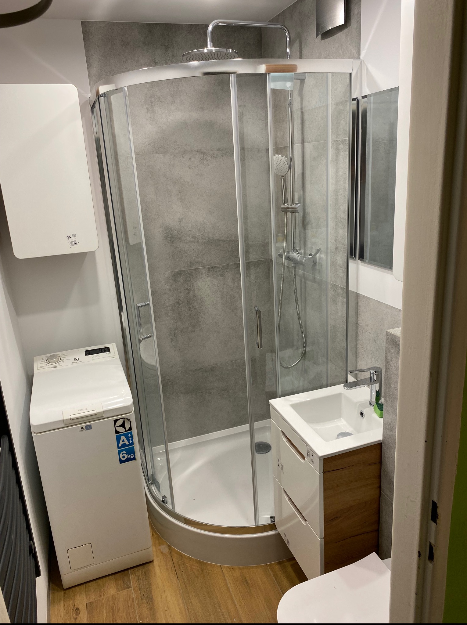 Nowoczesna łazienka po remoncie: kabina prysznicowa z deszczownicą, umywalka z szafką z elementami drewna, pralka. Szare płytki na ścianach, podłoga drewnopodobna.