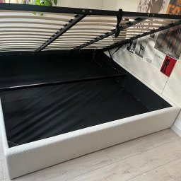 Damian Piekarski - Otwarta rama łóżka z pojemnikiem, biała tapicerka, czarne wnętrze, widoczne listwy stelaża. W tle plakaty na ścianie i podłoga z paneli.