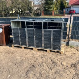 Damian Piekarski - Zmontowana, metalowa konstrukcja na palecie, prawdopodobnie obudowa na pojemniki na śmieci, stoi na ziemi obok brązowego pojemnika na odpady.