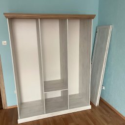 Damian Piekarski - Wnętrze pokoju z częściowo zmontowaną, biało-drewnianą szafą bez drzwi, opartą o błękitną ścianę. Obok leży jedna z drzwi szafy. Widoczna podłoga z paneli.