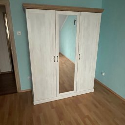 Damian Piekarski - Trzydrzwiowa, biała szafa z lustrem pośrodku, zwieńczona drewnianym blatem, stoi na drewnianej podłodze na tle turkusowej ściany. Widoczny fragment drzwi po lewej.