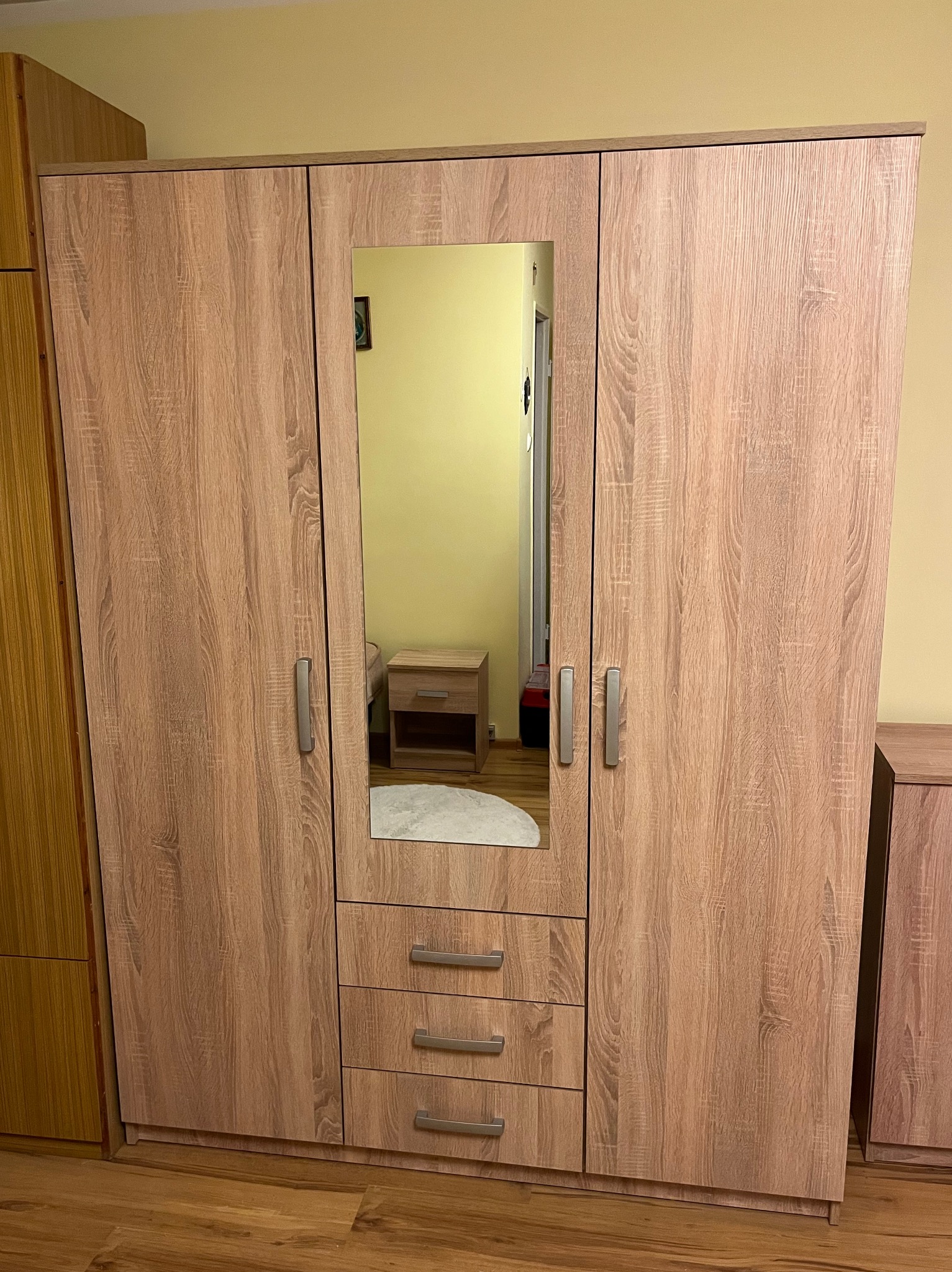 Trzydrzwiowa szafa z lustrem i szufladami w odcieniach drewna, widoczna na tle żółtej ściany i drewnianej podłogi. W lustrze odbija się fragment pokoju z szafką nocną.