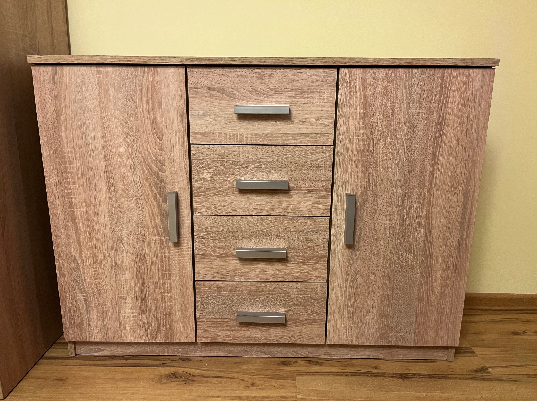 Zmontowana komoda z czterema szufladami i dwojgiem drzwi w odcieniach drewna, z metalowymi uchwytami, na tle żółtej ściany i drewnianej podłogi.