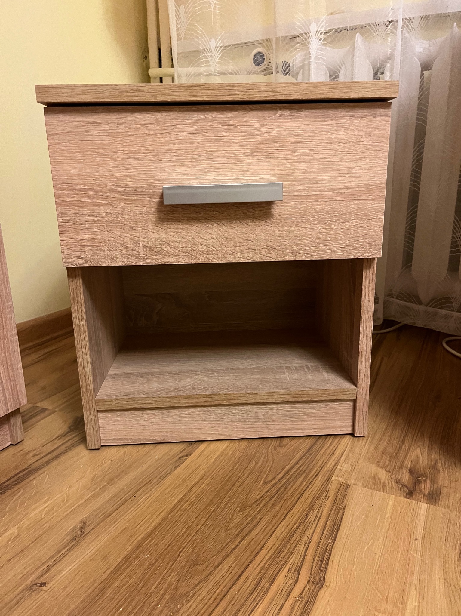 Drewniana szafka nocna z szufladą i otwartą półką, widoczna na tle parkietu i zasłony. Ujęcie z bliska, prezentujące detale wykończenia.