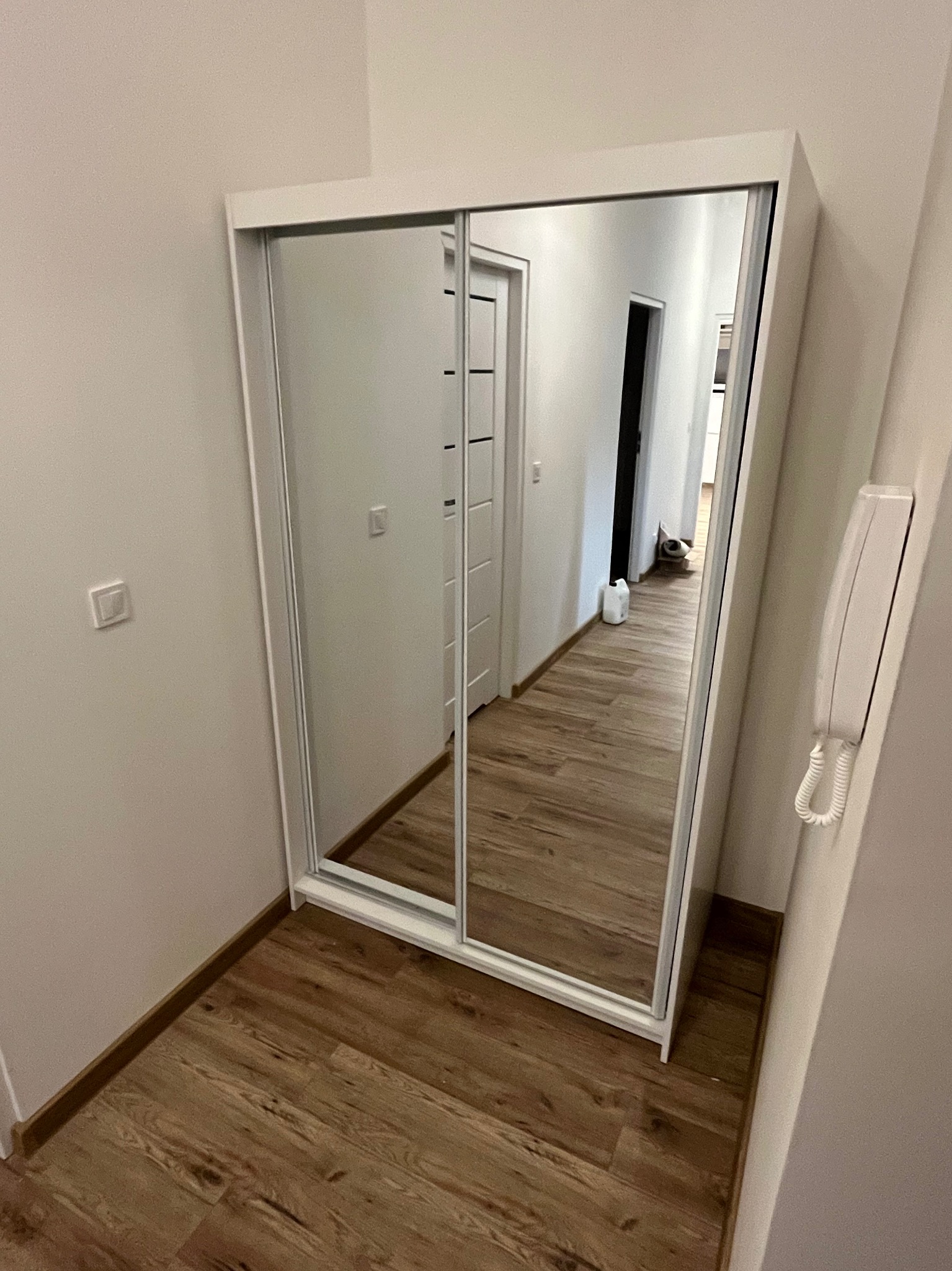 Biała szafa przesuwna z lustrami w przedpokoju, odbijająca wnętrze mieszkania. Widoczne panele podłogowe i domofon na ścianie. Minimalistyczny design.