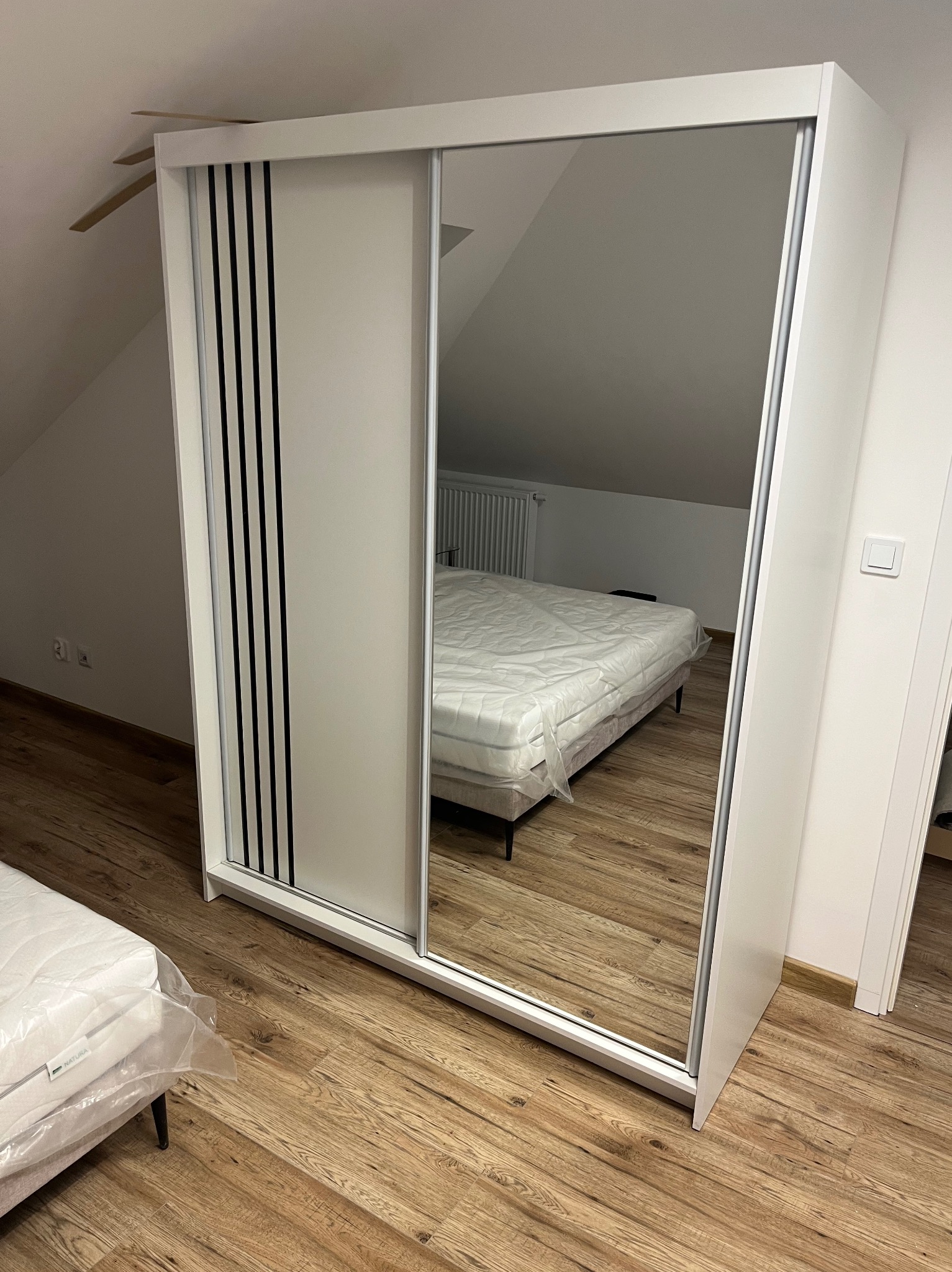 Biała szafa przesuwna z lustrem, odbijająca łóżko z materacem w folii, na drewnianej podłodze. Ukośny sufit, minimalistyczny design.