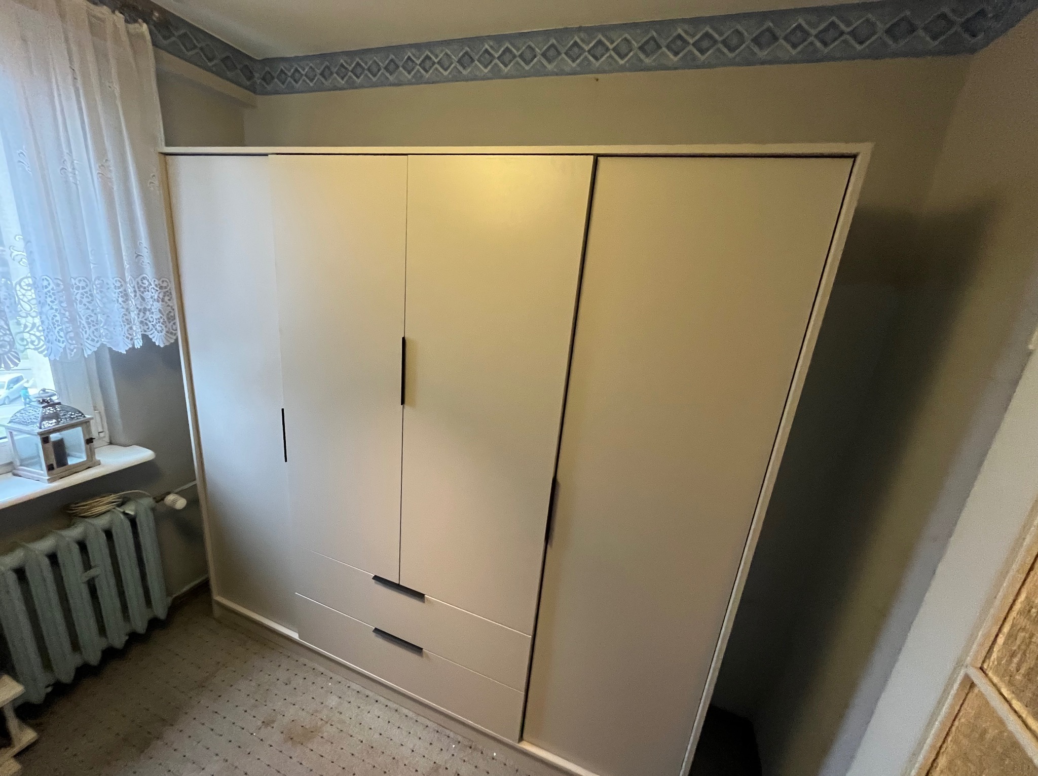 Jasna, trzydrzwiowa szafa z szufladami w Gołyminie-Ośrodku, ustawiona w rogu pokoju. Widoczny grzejnik i okno z dekoracyjną lampką. Minimalistyczny design.