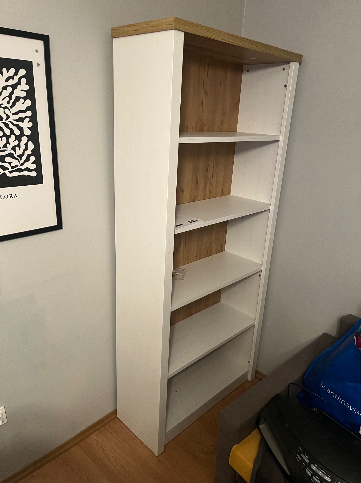 Wysoki, biały regał z drewnianym blatem i półkami, ustawiony w rogu pokoju. Na ścianie wisi czarno-biały plakat w ramie. Widoczna podłoga z paneli.