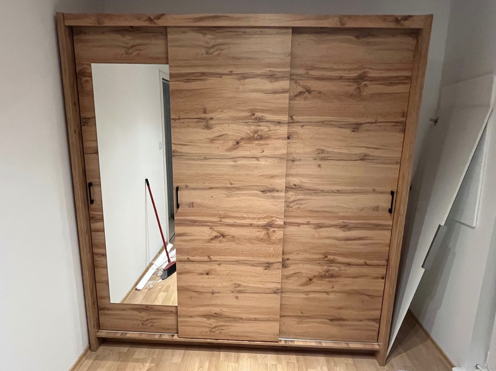 Drewniana szafa przesuwna z lustrem, widoczna w częściowym ujęciu. W lustrze odbija się fragment pomieszczenia z miotłą. Ustawiona na podłodze z paneli.