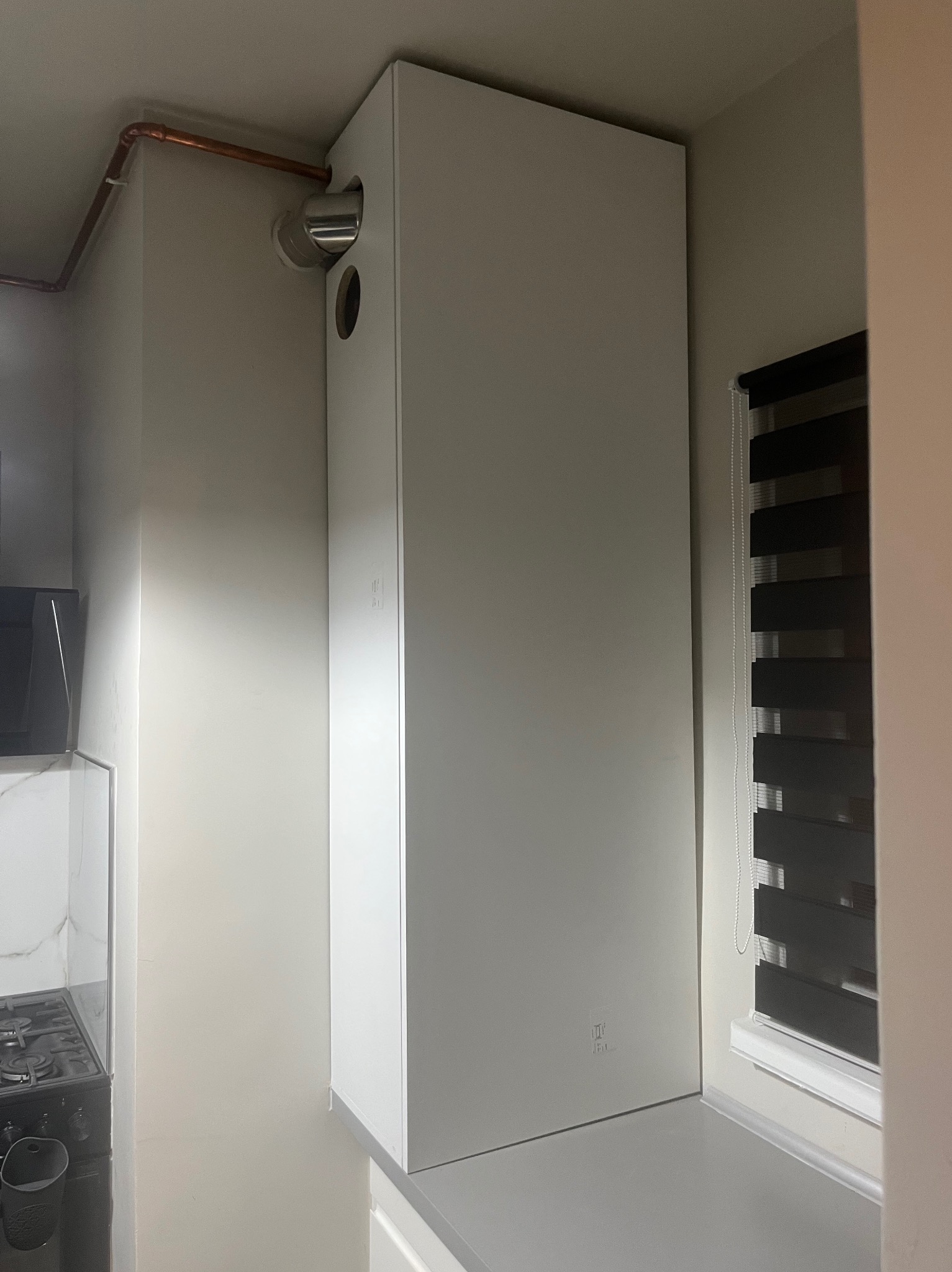 Biała zabudowa urządzenia z miedzianą rurą w Gołymin-Ośrodek. Minimalistyczny design, widoczne przyłącza. Estetyczne wykończenie w jasnym wnętrzu.