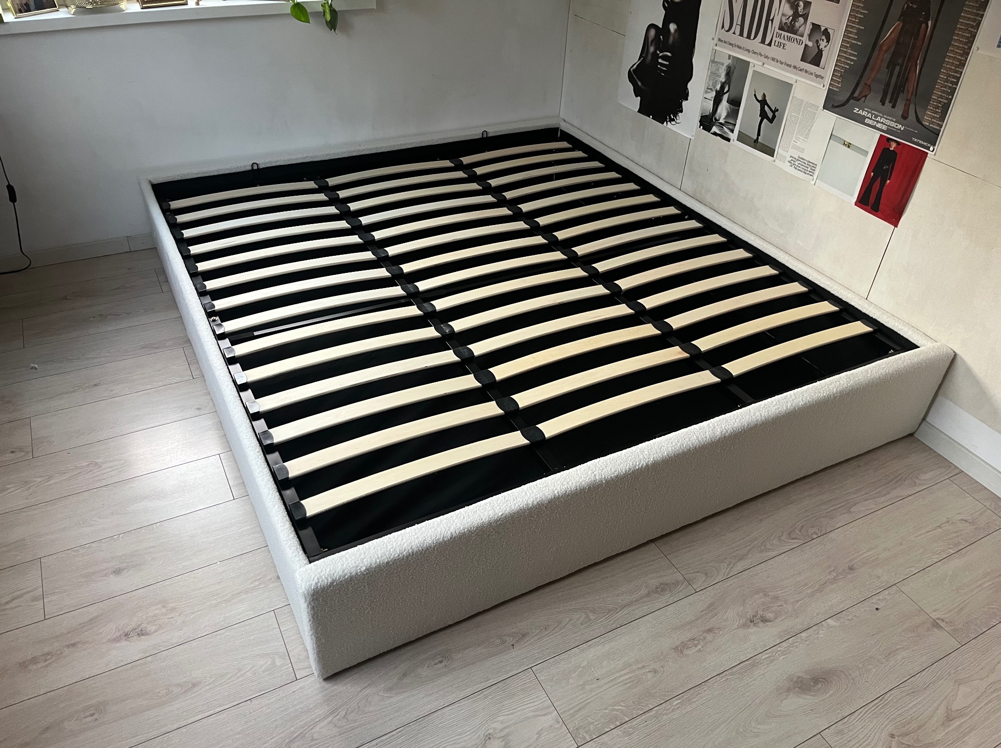 Biała rama łóżka z drewnianym stelażem listwowym na podłodze z paneli. Na ścianie plakaty w czarno-białej tonacji. Widok z góry, ukośnie.