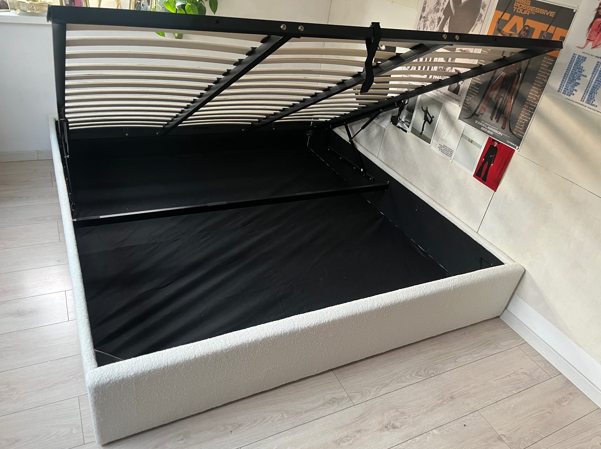 Otwarta rama łóżka z pojemnikiem, biała tapicerka, czarne wnętrze, widoczne listwy stelaża. W tle plakaty na ścianie i podłoga z paneli.