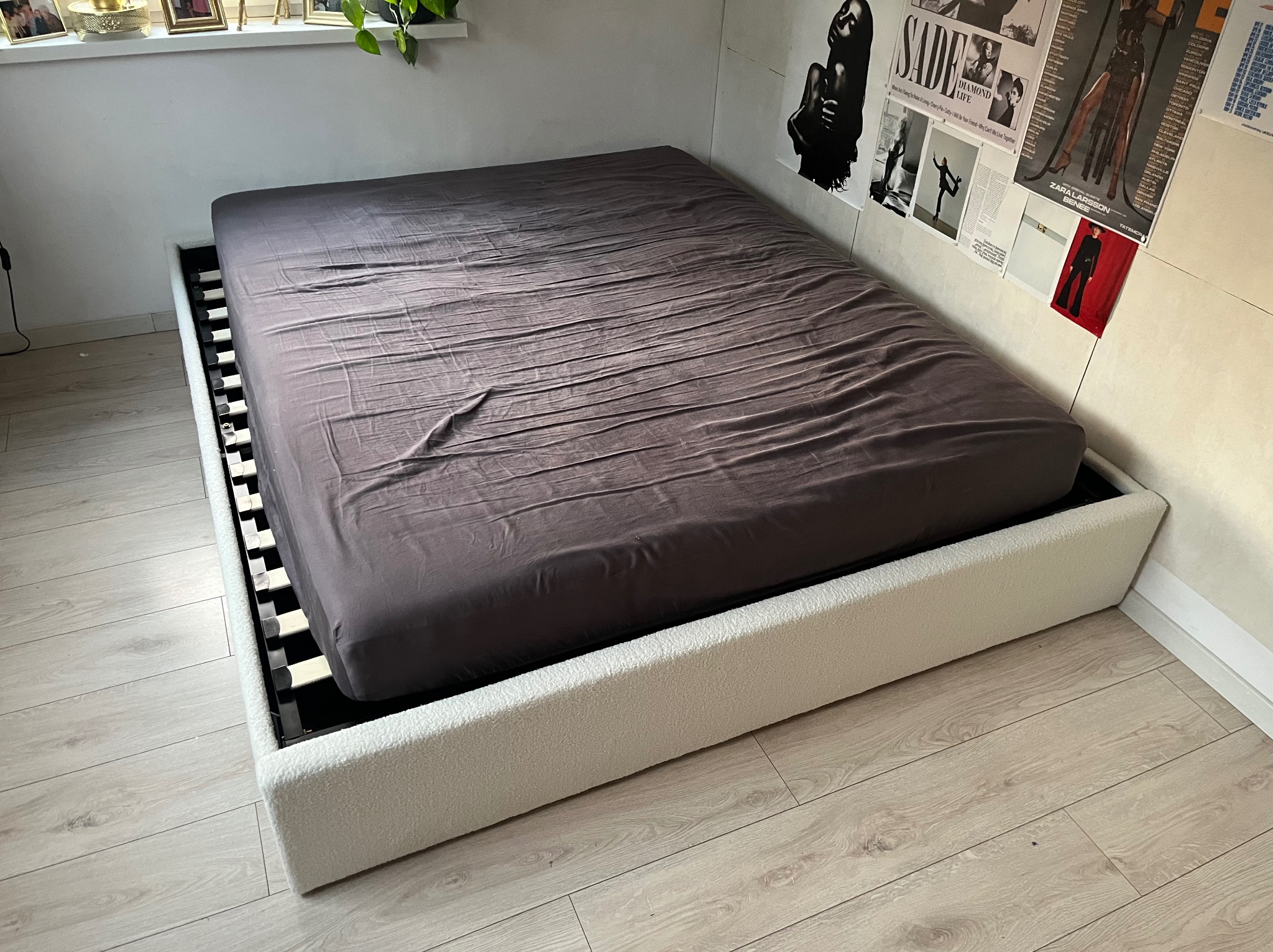 Nowoczesne łóżko z tapicerowanym, kremowym obiciem i szarą pościelą, ustawione na drewnianej podłodze. Na ścianie plakaty w stylu vintage.