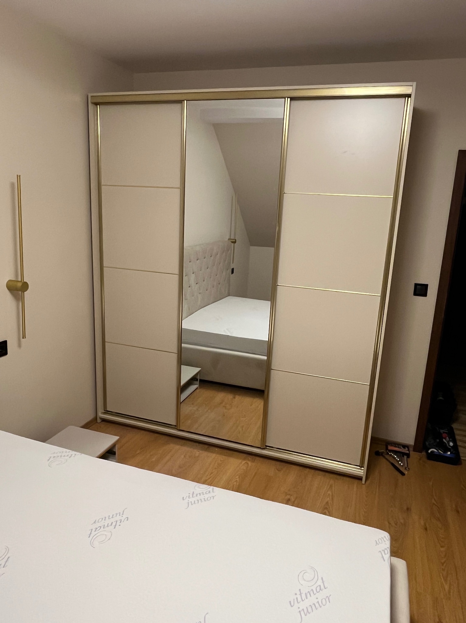 Beżowa szafa przesuwna z lustrem w sypialni, widoczny fragment łóżka i lampka nocna. Na podłodze leżą narzędzia.