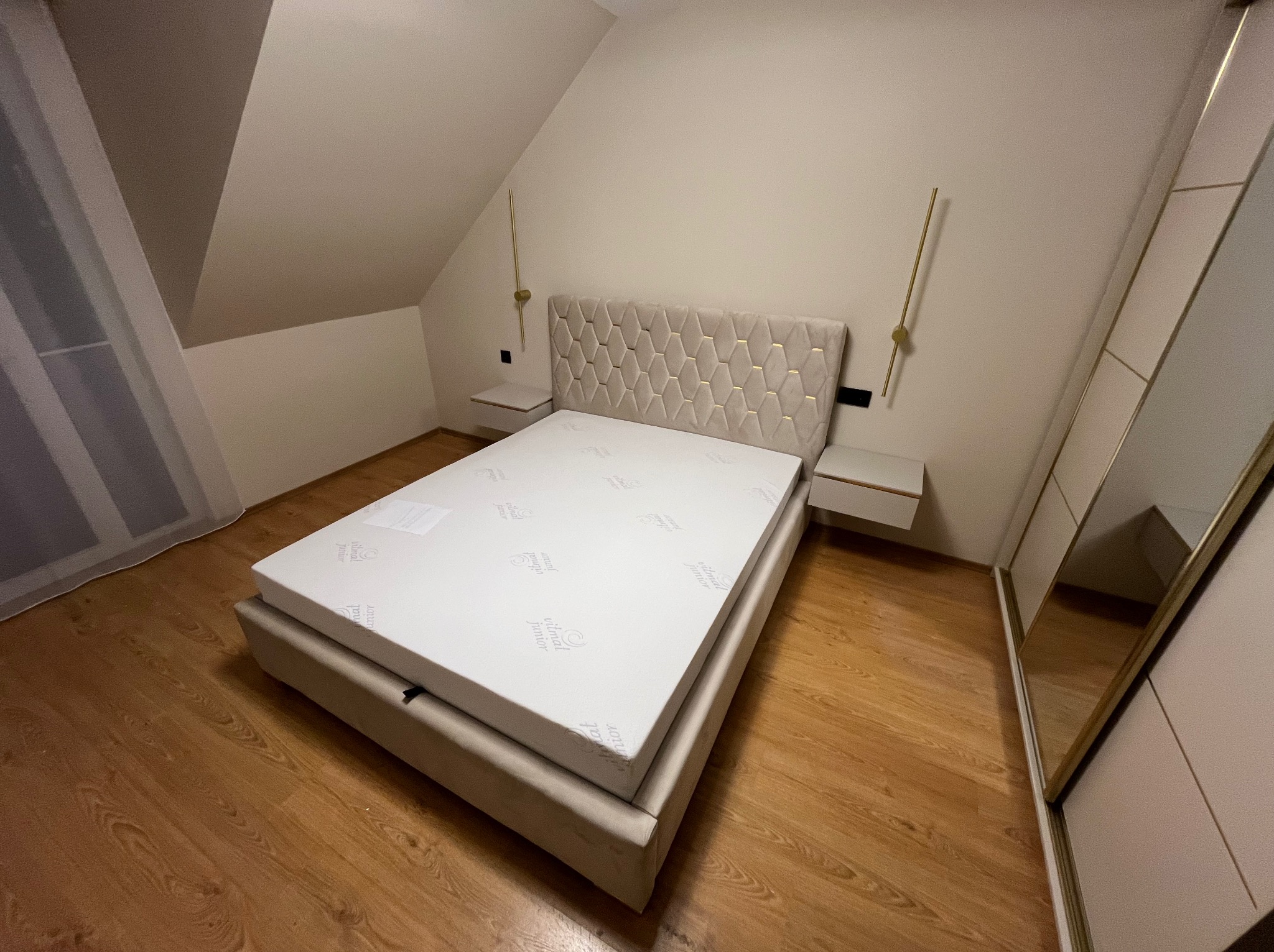 Beżowe łóżko z tapicerowanym zagłówkiem w romby, z białym materacem w sypialni na poddaszu. Po bokach łóżka szafki nocne i złote lampki.