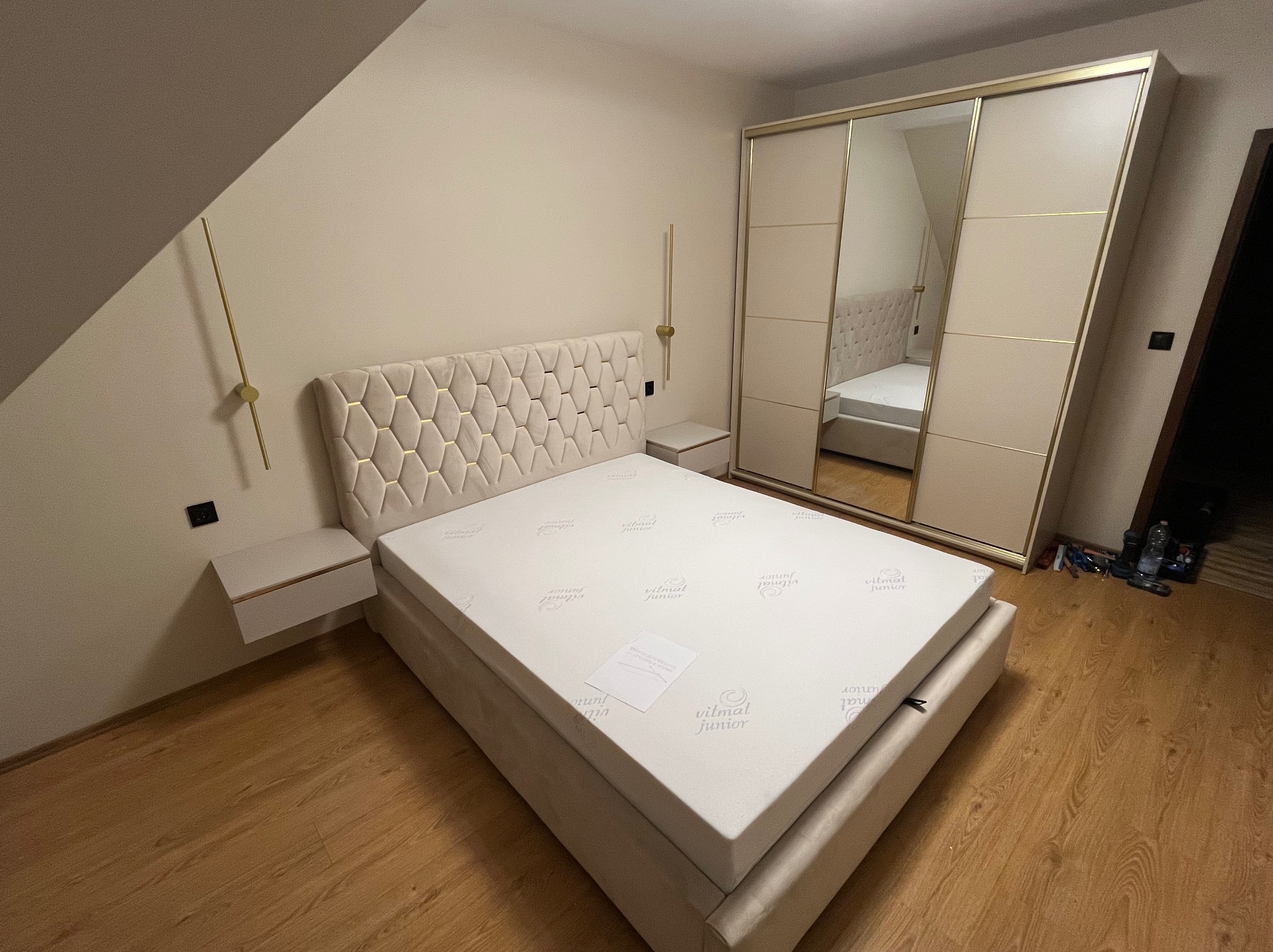 Beżowe łóżko z geometrycznym zagłówkiem i szafki nocne w komplecie, ustawione w sypialni z drewnianą podłogą i szafą z lustrzanymi drzwiami przesuwnymi.