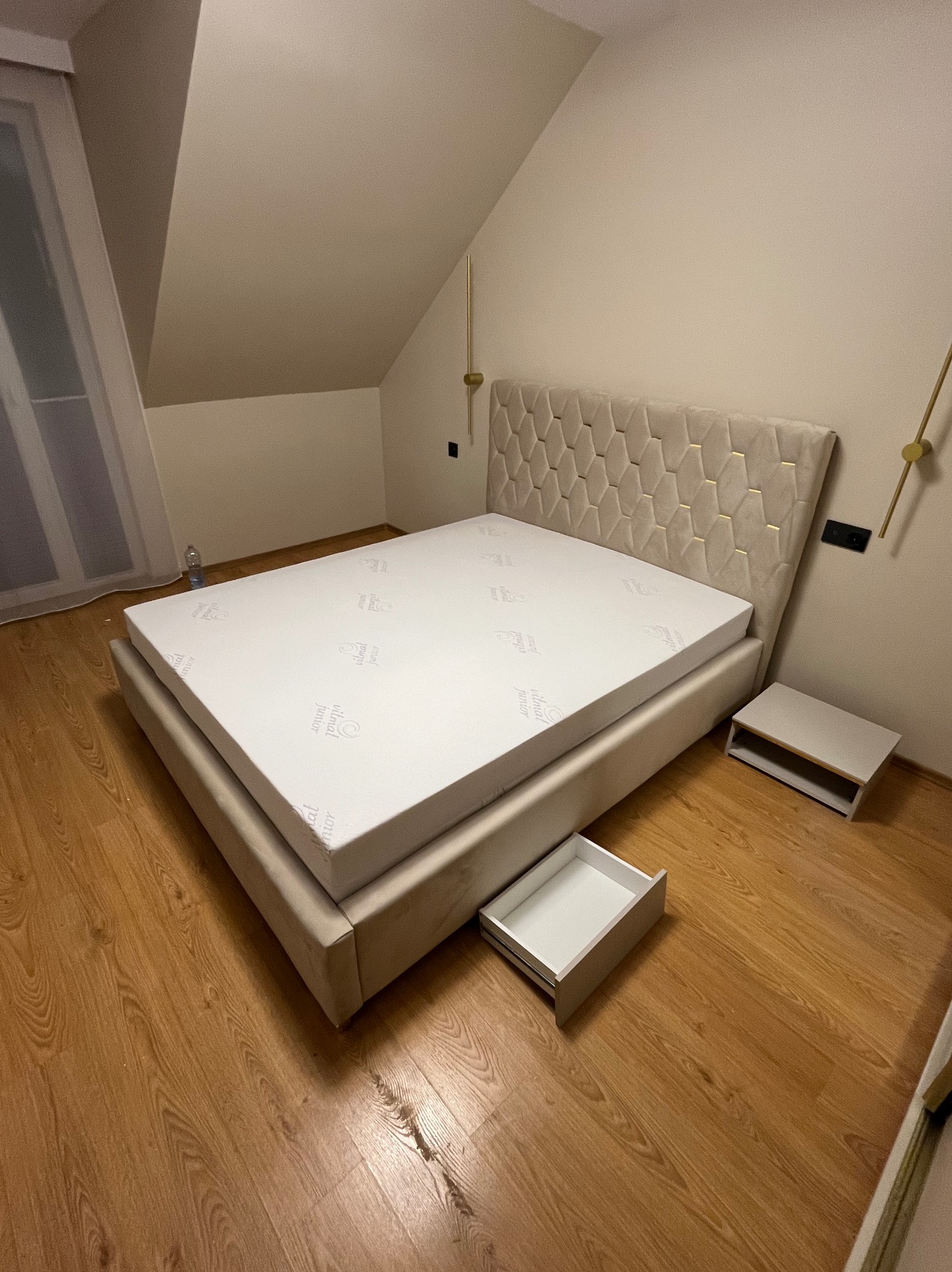 Beżowe łóżko z tapicerowanym zagłówkiem w geometryczne wzory, wysunięta szuflada, drewniana podłoga. Nowoczesny design w sypialni na poddaszu.