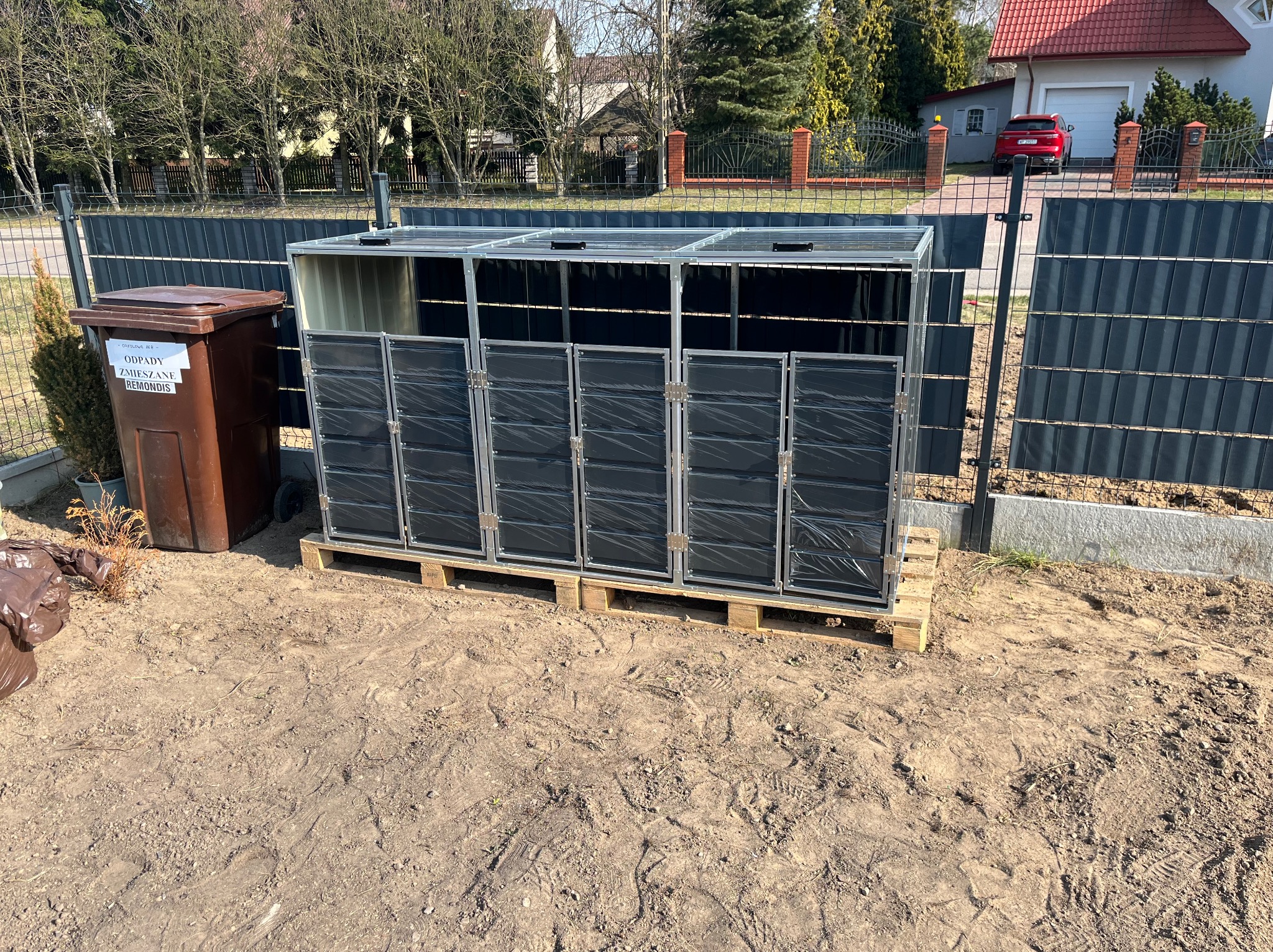 Zmontowana, metalowa konstrukcja na palecie, prawdopodobnie obudowa na pojemniki na śmieci, stoi na ziemi obok brązowego pojemnika na odpady.