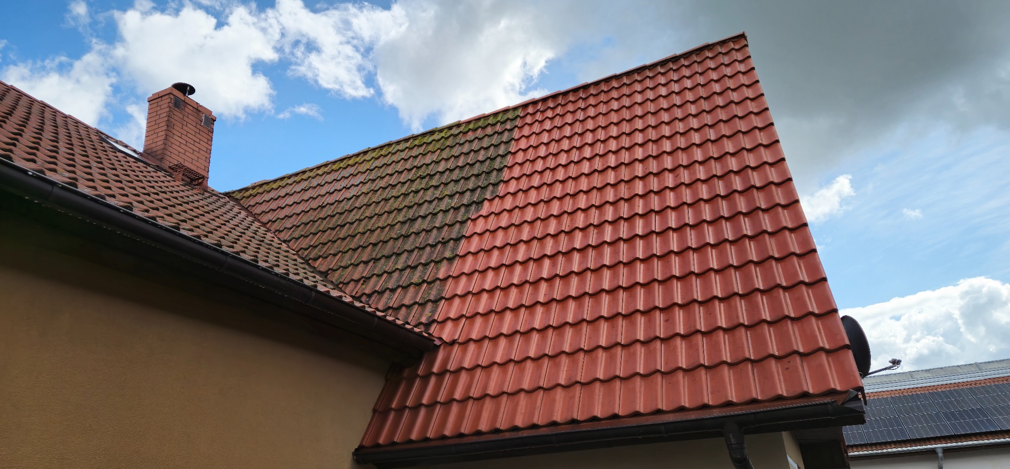Częściowo wyczyszczony dach z czerwonej dachówki, widoczna różnica między brudną a czystą powierzchnią. Kominy, rynna i fragment elewacji w kadrze.