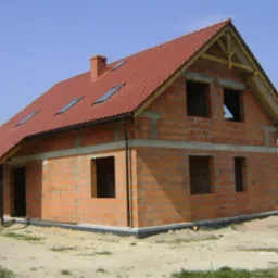 Surowy, dwukondygnacyjny dom z czerwonej cegły z dachem z dachówki, widoczne okna dachowe, w stanie budowy, otoczony piaszczystym terenem i zielenią.