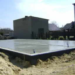 Świeżo wylana płyta fundamentowa z hydroizolacją, otoczona piaskiem i ziemią. W tle budynki mieszkalne i błękitne niebo. Widoczne detale jak poziomice i fragmenty szalunku.