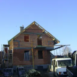 Budowa domu z cegły w Tarnowskich Górach. Widoczne rusztowania, częściowo wymurowane ściany i konstrukcja dachu w trakcie montażu. Na pierwszym planie samochód dostawczy.