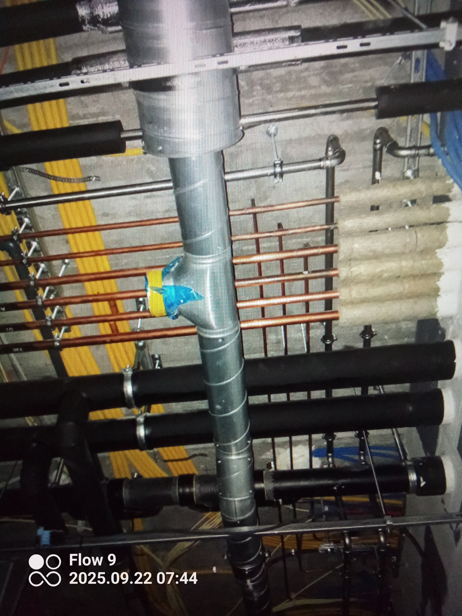 Widok na sufit z licznymi rurami instalacji: miedziane rury grzewcze, czarne rury izolowane i srebrne rury wentylacyjne. Widoczne mocowania i elementy instalacyjne.