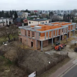 Dwukondygnacyjny budynek w stanie surowym otwartym z betonu i cegły. Na budowie stoi pomarańczowa ładowarka teleskopowa. W tle zabudowa miejska.