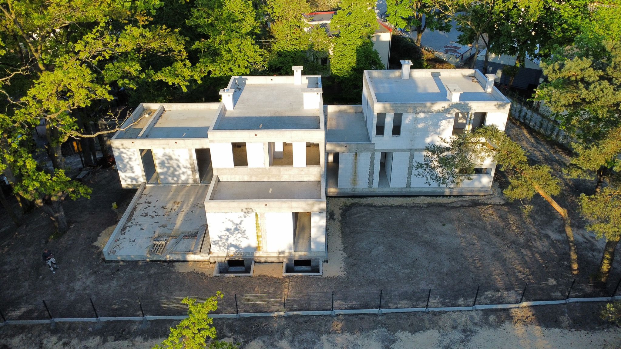 Surowy stan dwukondygnacyjnego domu z białego betonu komórkowego, widok z góry, otoczonego zielenią drzew i ogrodzeniem z siatki. Widoczny zarys przyszłych okien.