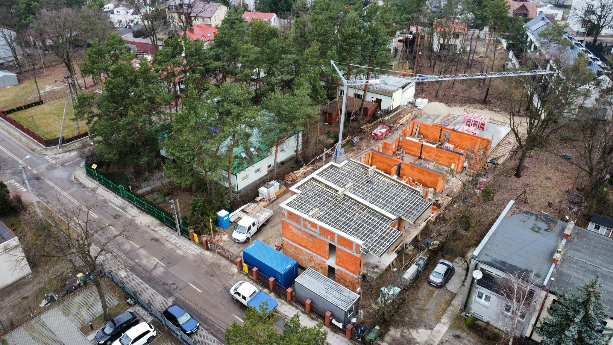 Z lotu ptaka: budowa domów z czerwonej cegły, widoczne ściany, strop zbrojony, dźwig budowlany. W tle drzewa i zabudowa mieszkalna.