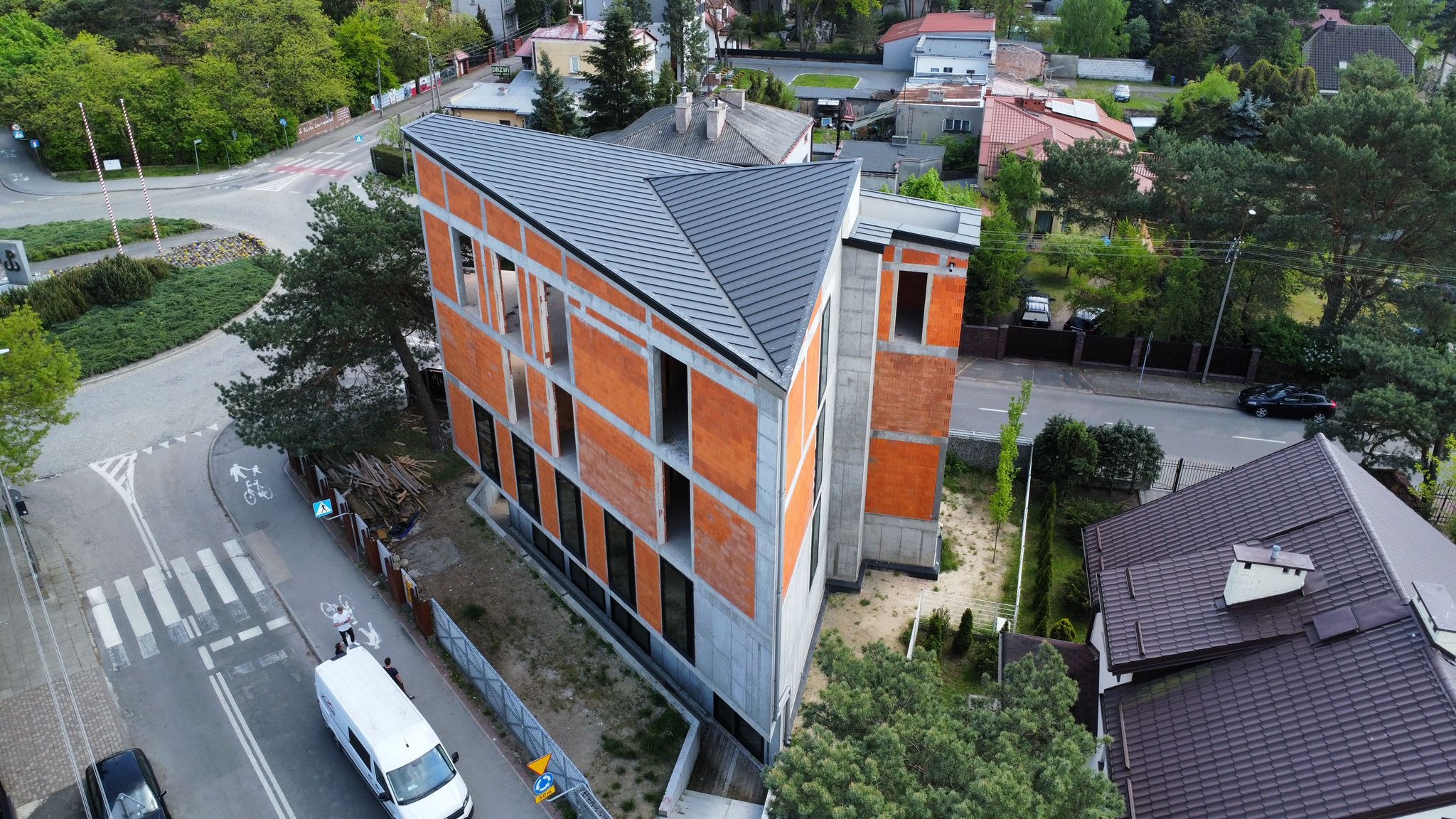 Surowy, dwukondygnacyjny budynek z cegły i betonu, z zamontowanym, grafitowym dachem, widziany z lotu ptaka. Okna bez szyb, otoczenie w trakcie zagospodarowania.