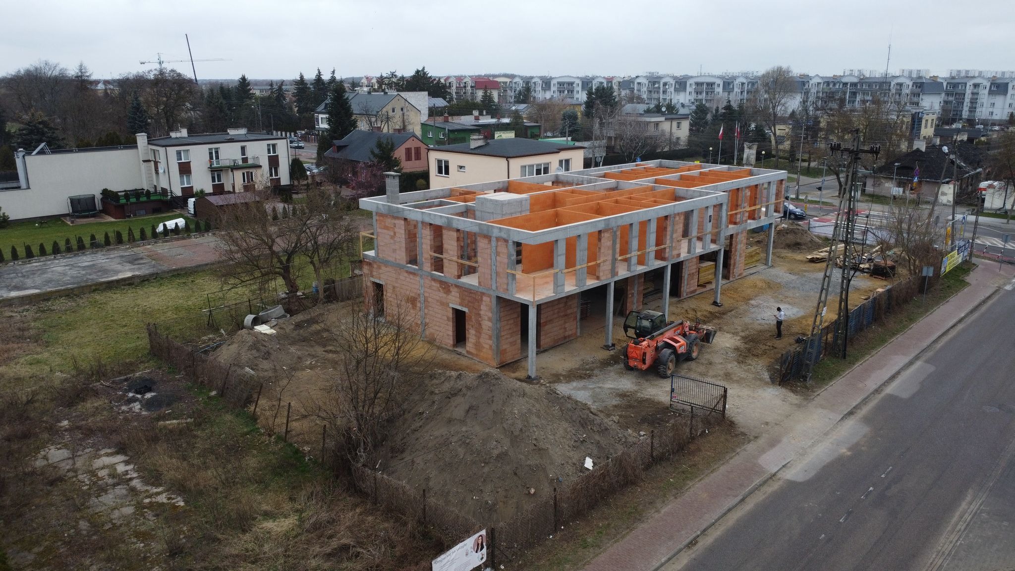 Dwukondygnacyjny budynek w stanie surowym otwartym z betonu i cegły. Na budowie stoi pomarańczowa ładowarka teleskopowa. W tle zabudowa miejska.