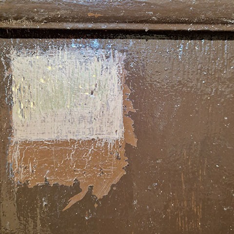 Fragment odnawianej, brązowej, drewnianej powierzchni. Widoczny prostokąt z nałożoną białą farbą, częściowo odsłaniający oryginalną warstwę. Detal procesu konserwacji.