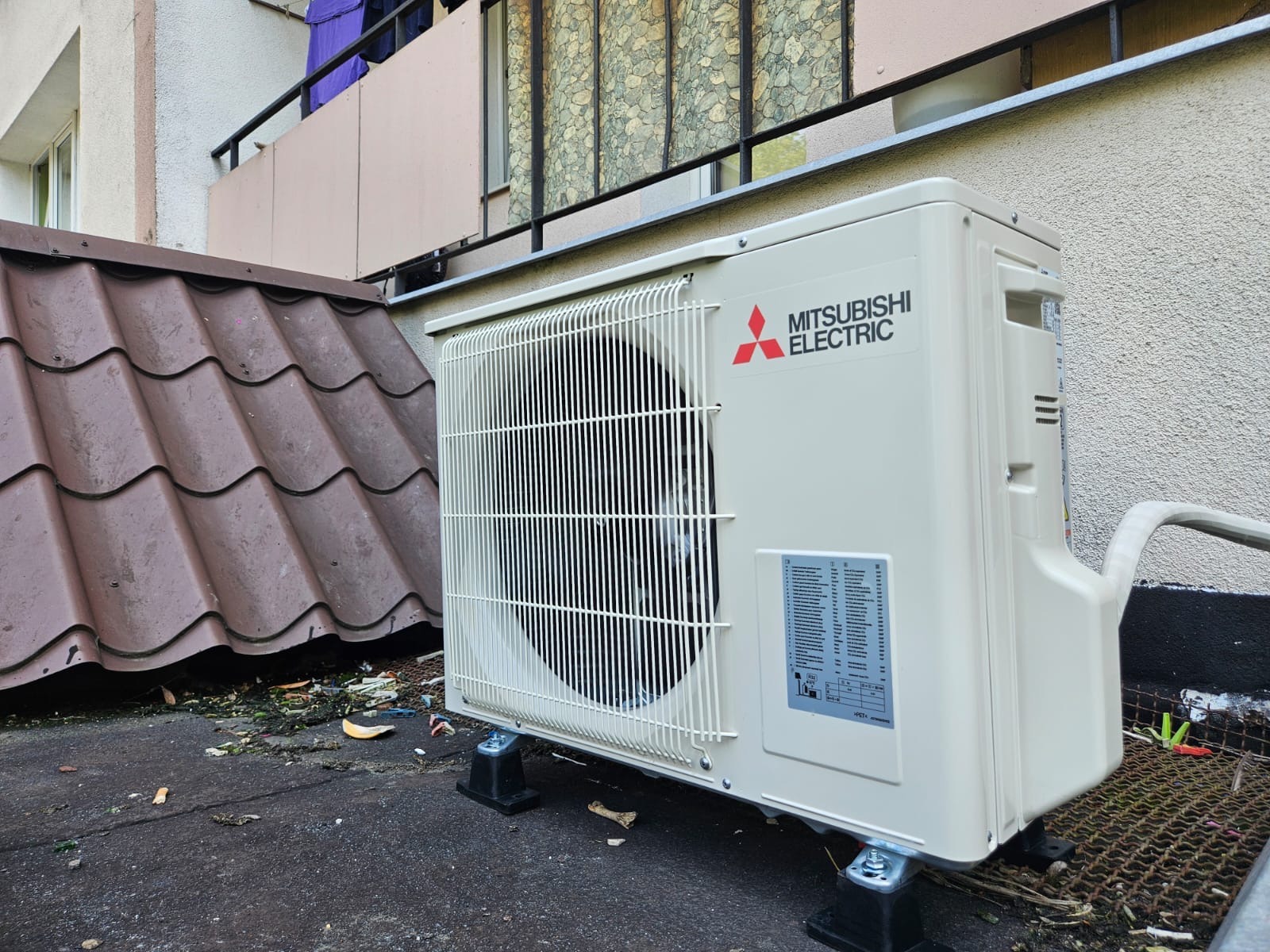 Zamontowana jednostka zewnętrzna klimatyzacji Mitsubishi Electric na dachu budynku, widoczna instalacja, tło stanowi elewacja i balkon.