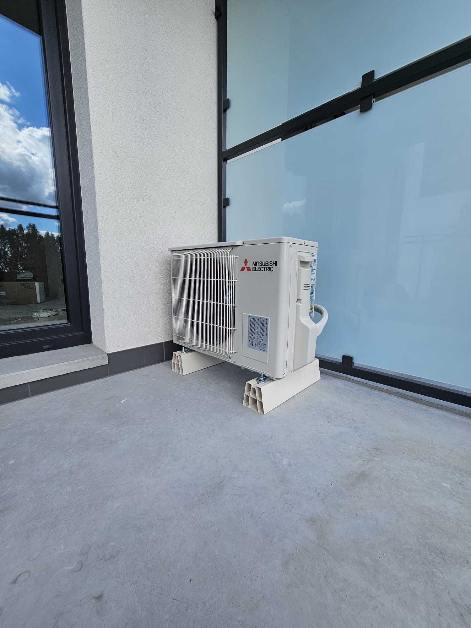 Zewnętrzna jednostka klimatyzacji Mitsubishi Electric zamontowana na betonowym podłożu, z widocznym oknem i szklaną ścianą w tle.