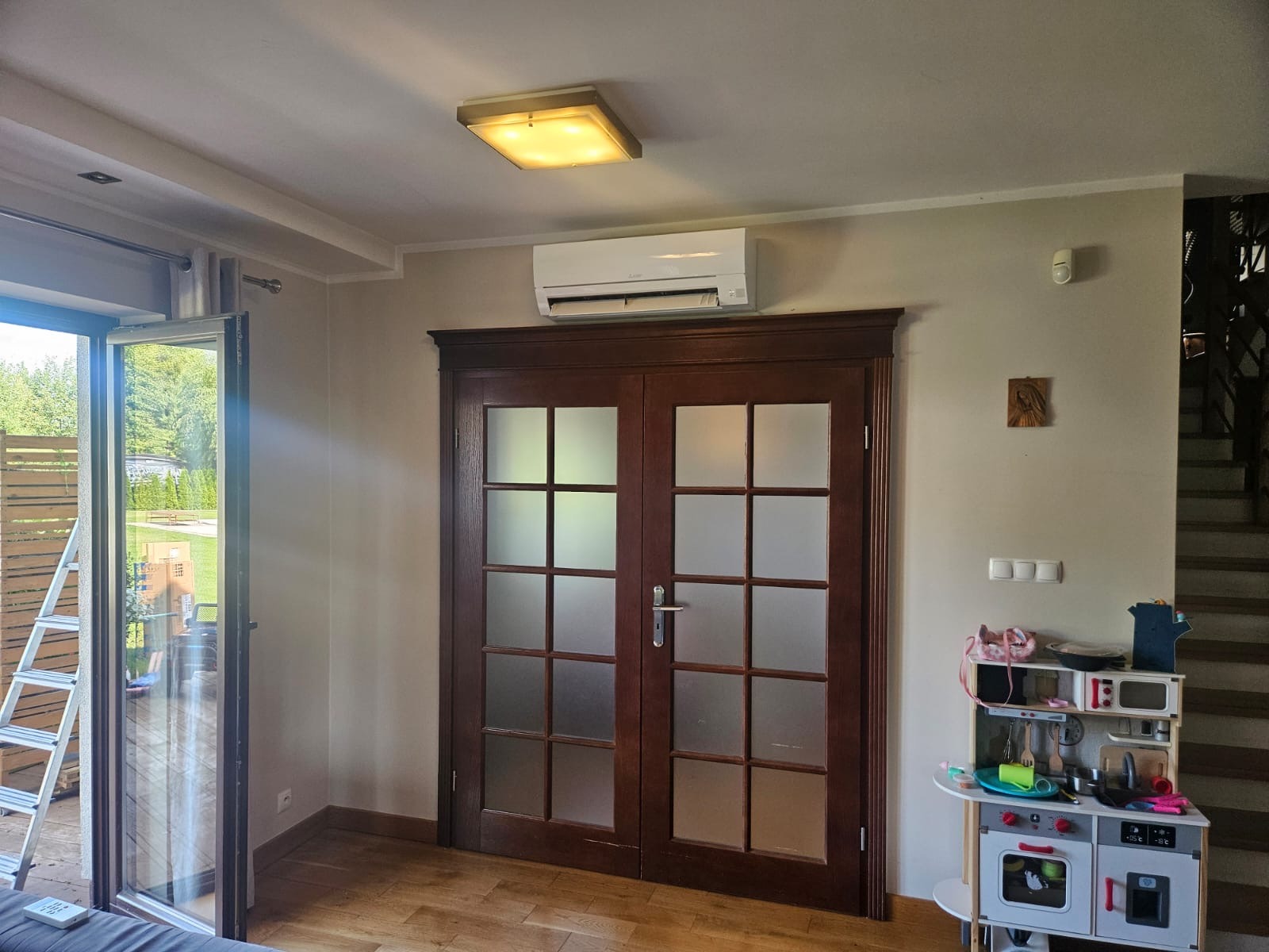 Biała klimatyzacja ścienna nad drewnianymi drzwiami z szybami, widok na wnętrze domu z zabawkami i schodami.