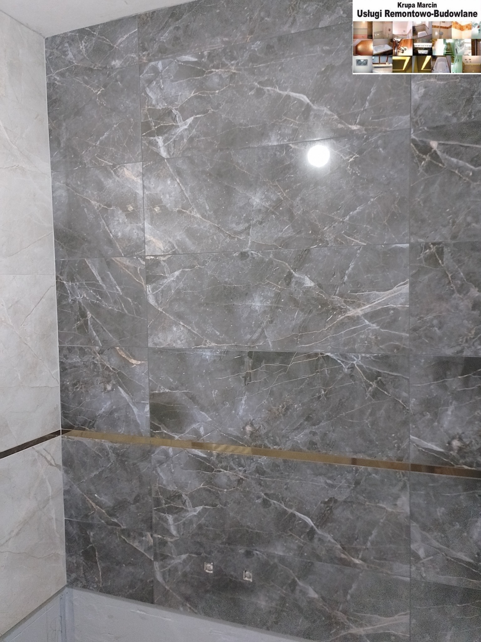 Ściana wyłożona szarymi płytkami imitującymi marmur, z poziomym złotym paskiem dekoracyjnym. Fragment narożnika w łazience, widoczne fugi i oświetlenie.