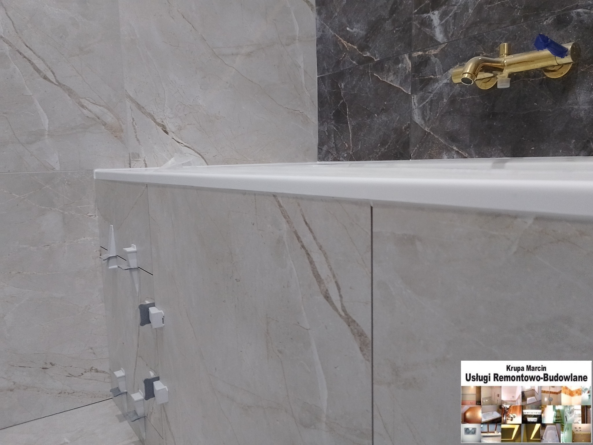 Fragment wykończonej łazienki z beżowymi płytkami imitującymi marmur, złotą armaturą i detalami montażowymi. Widoczny fragment wanny.