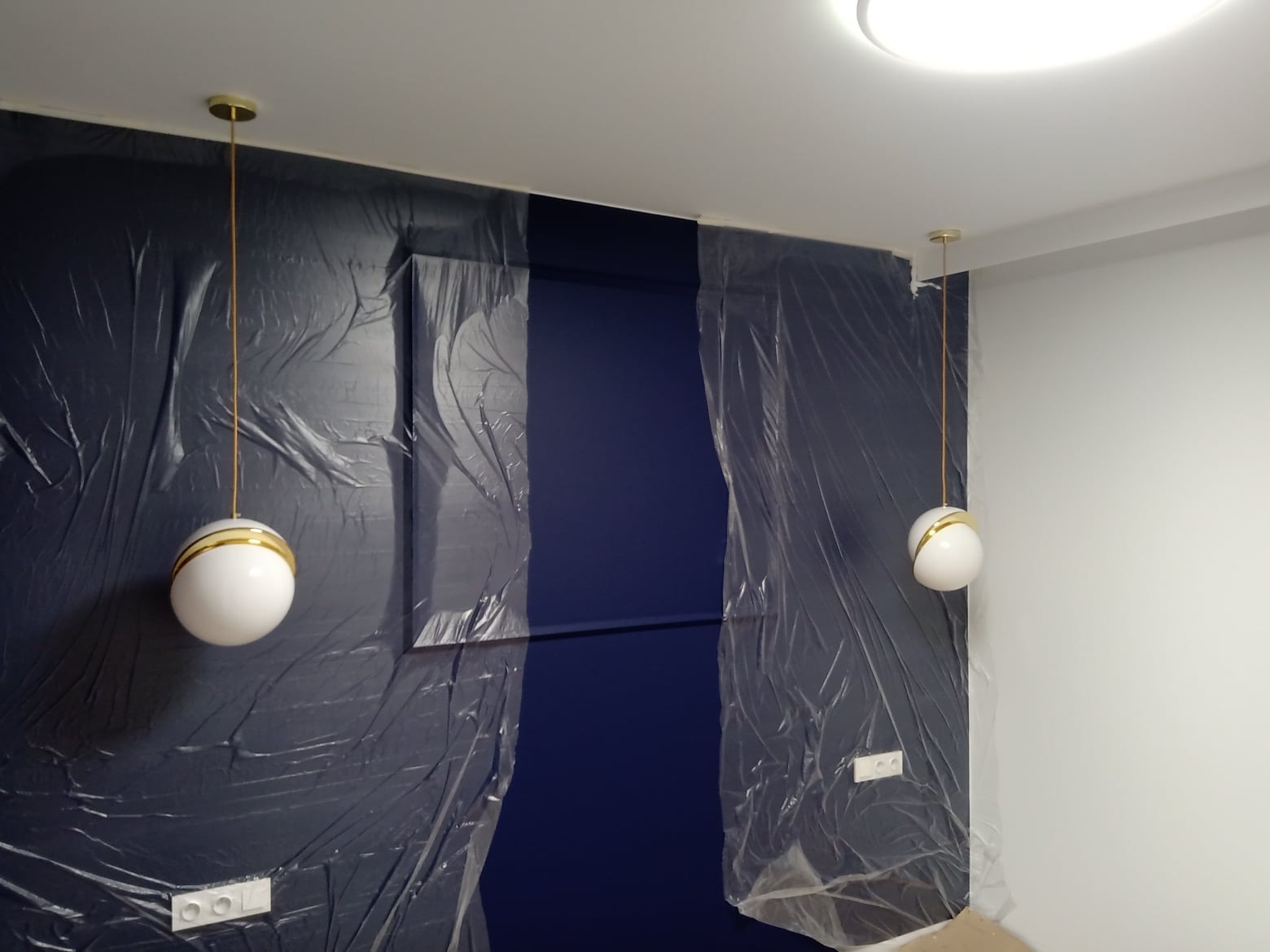 Przygotowanie do malowania: zabezpieczona folią ściana z oświetleniem, fragment pomalowanej ściany w odcieniu granatu, widoczne gniazdka elektryczne.