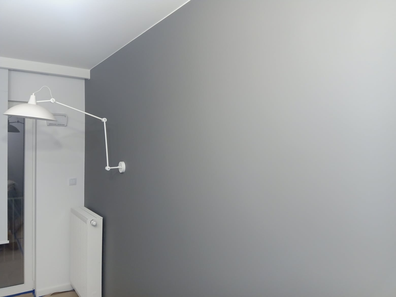 Ściana pomalowana na szaro z białym, regulowanym kinkietem i grzejnikiem. Widok z bliska, równomierne krycie farby, stonowana kolorystyka, nowoczesny design.