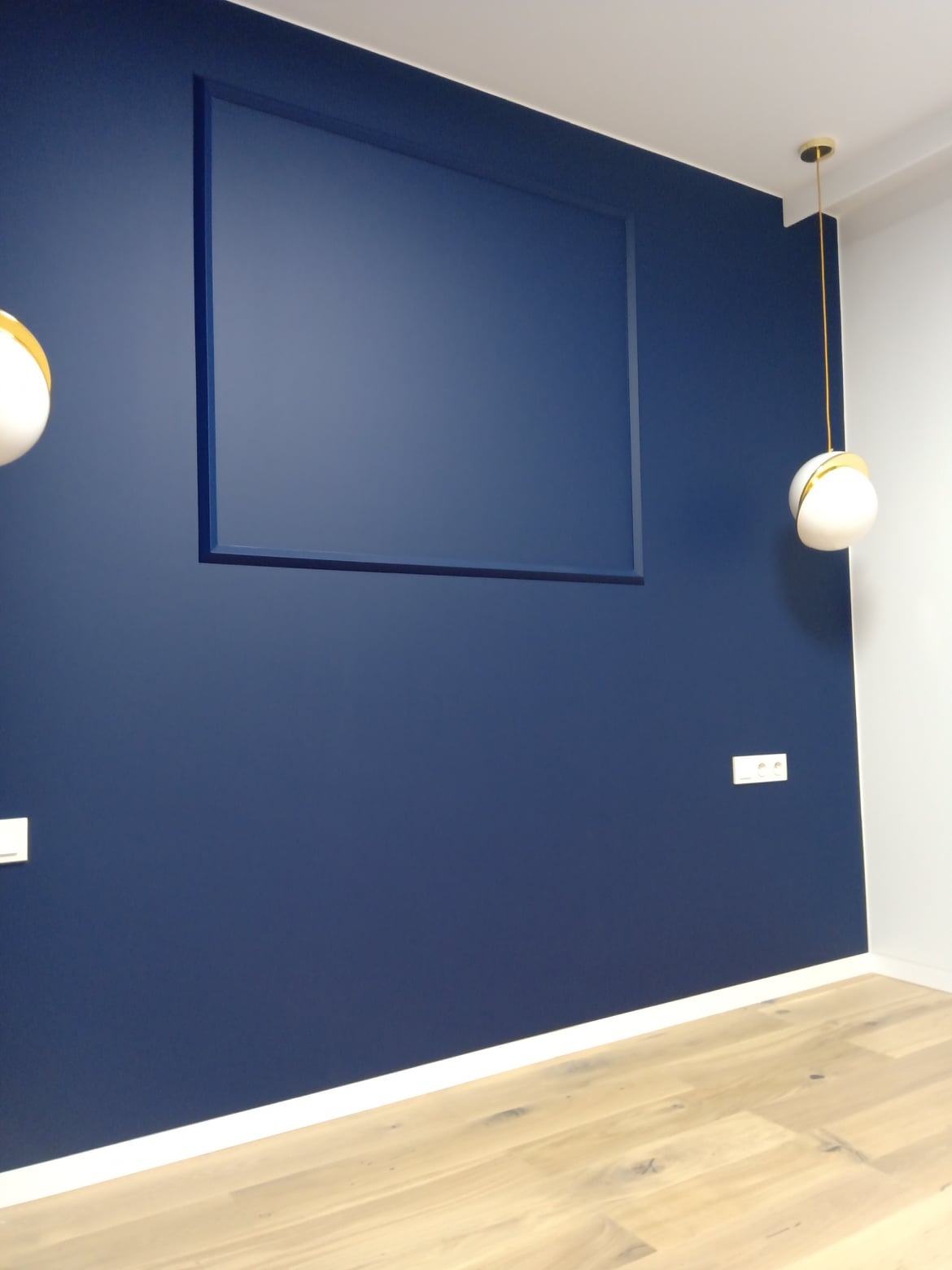 Granatowa ściana z dekoracyjną ramą w kolorze ściany, oświetlona nowoczesnymi lampami. Widoczne gniazdka elektryczne i drewniana podłoga.