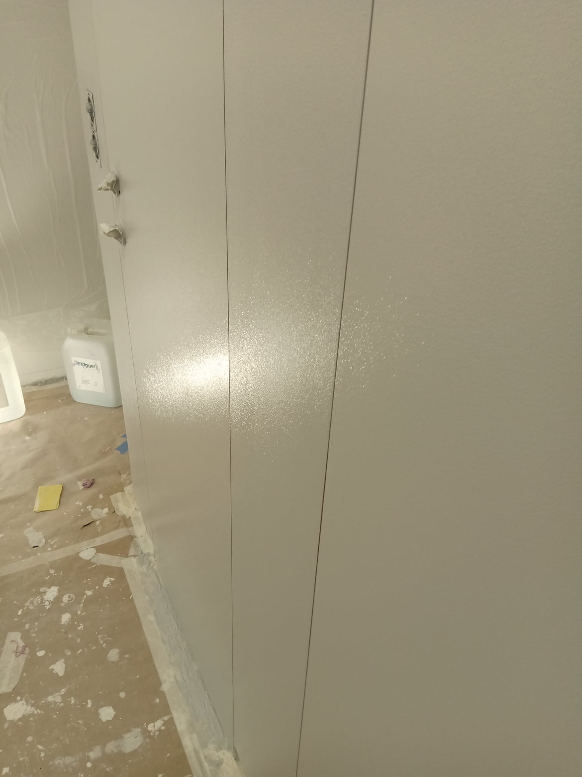 Świeżo pomalowana, jasnoszara ściana z widocznym połyskiem, zabezpieczona folią i taśmą malarską przy podłodze, z pojemnikiem farby w tle.