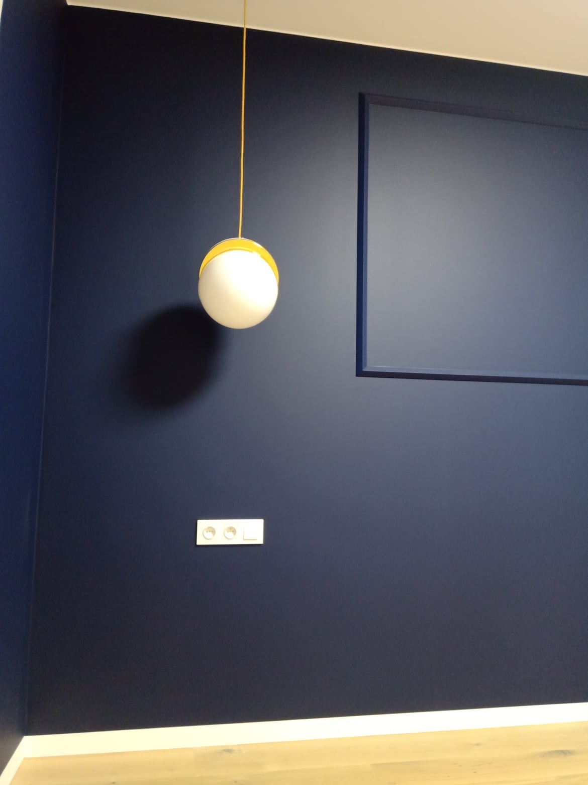 Granatowa ściana z gładzią, dekoracyjną ramką i designerską lampą wiszącą. Widoczny kontakt elektryczny i fragment podłogi.
