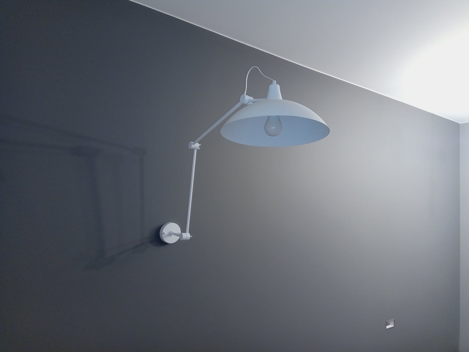 Ściana w odcieniach szarości z zamontowaną białą lampą, oświetlającą wnętrze. Minimalistyczny design, gładka powierzchnia ściany, nowoczesny styl.