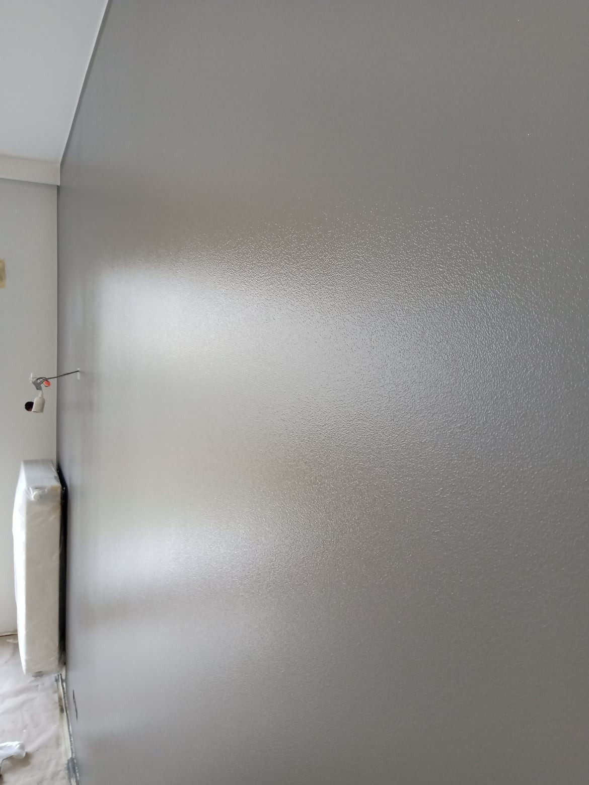 Świeżo pomalowana, szara ściana w pokoju, widoczny grzejnik w folii ochronnej i gładka powierzchnia ściany. Nowoczesne wykończenie wnętrza.