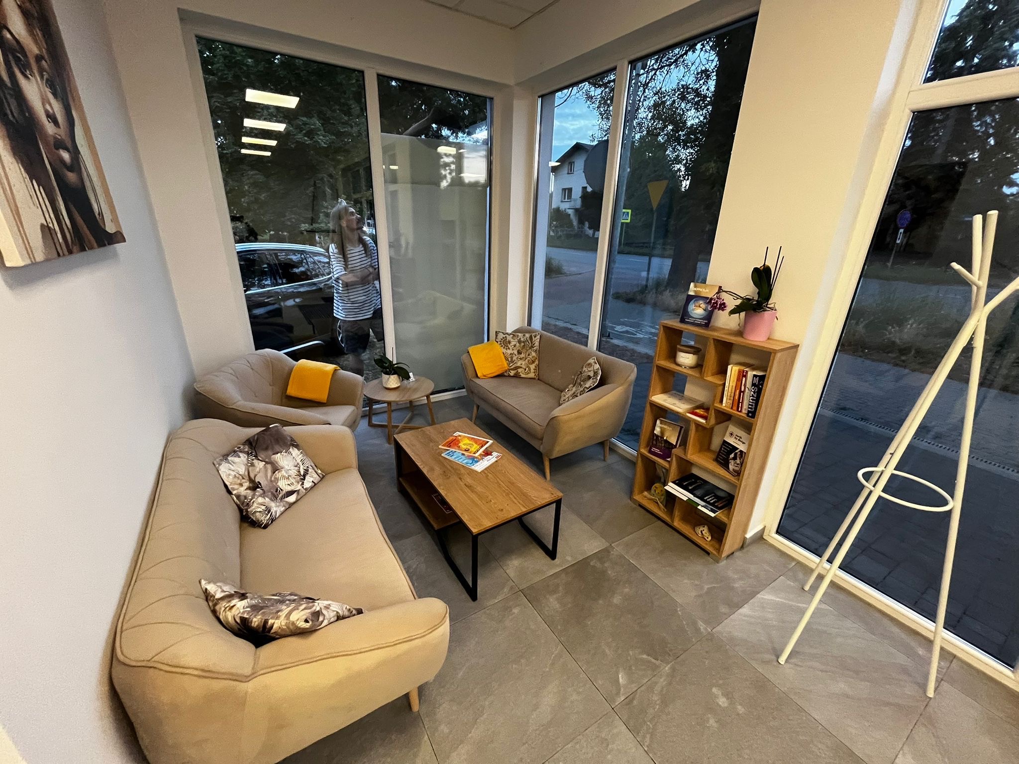 Jasne wnętrze poczekalni z beżowymi sofami, stolikiem kawowym i regałem na książki. Za oknem widoczna osoba i zieleń. Obraz na ścianie dodaje charakteru.